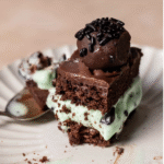 A slice of mint chocolate chip ice cream cake topped with chocolate ganache and a truffle on a white plate