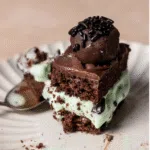 A slice of mint chocolate chip ice cream cake topped with chocolate ganache and a truffle on a white plate